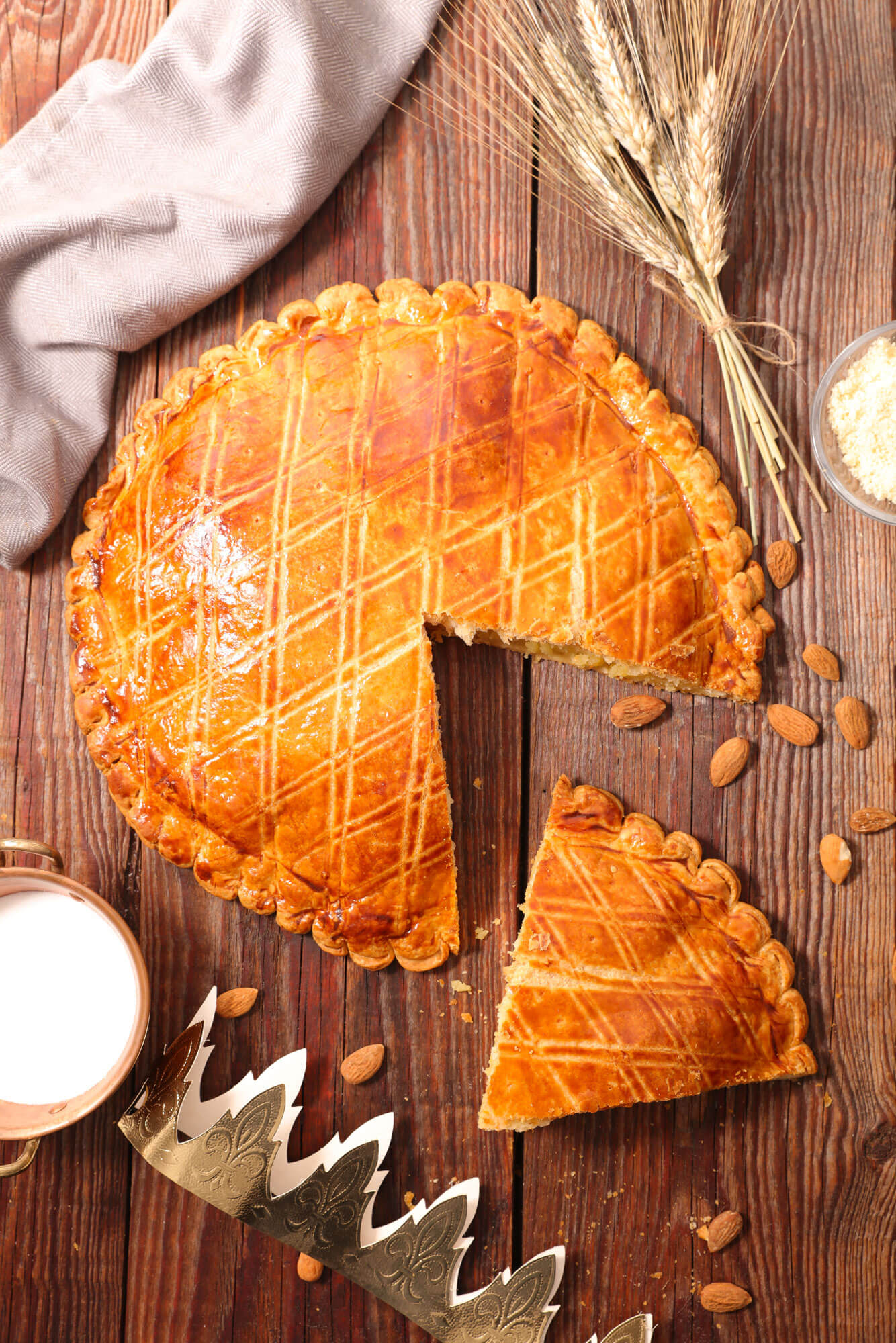 Gros plan sur une galette vue du dessus avec part découpée, amandes dispersées, épi de blé posés sur une table en bois