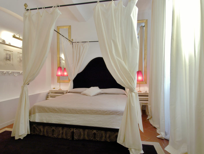 Grand lit avec baldaquins sur les 4 côtés, et tête de lit noire accompagnée de miroirs et lampes de chevets pour une ambiance romantique type Saint Valentin