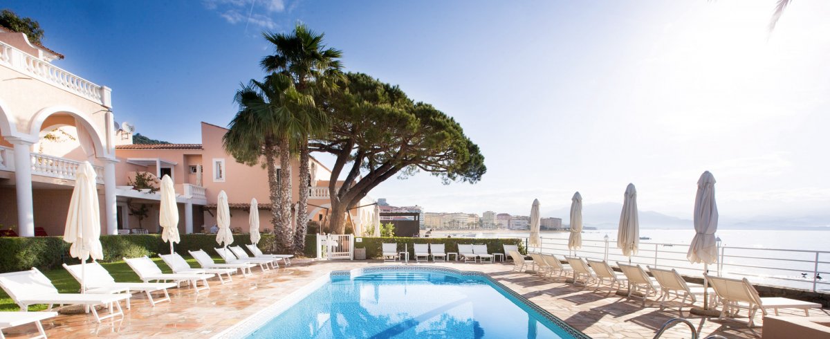 Paysage côtier de Corse, avec vaste piscine extérieure entourée de transats et de parasols.