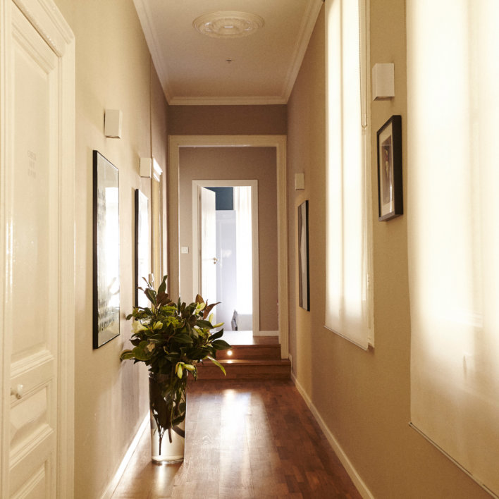 Couloir de style moderne dans des couleurs claires avec parquet au sol décoré de cadres au murs, avec un grand vase fleuri posé à même le sol.