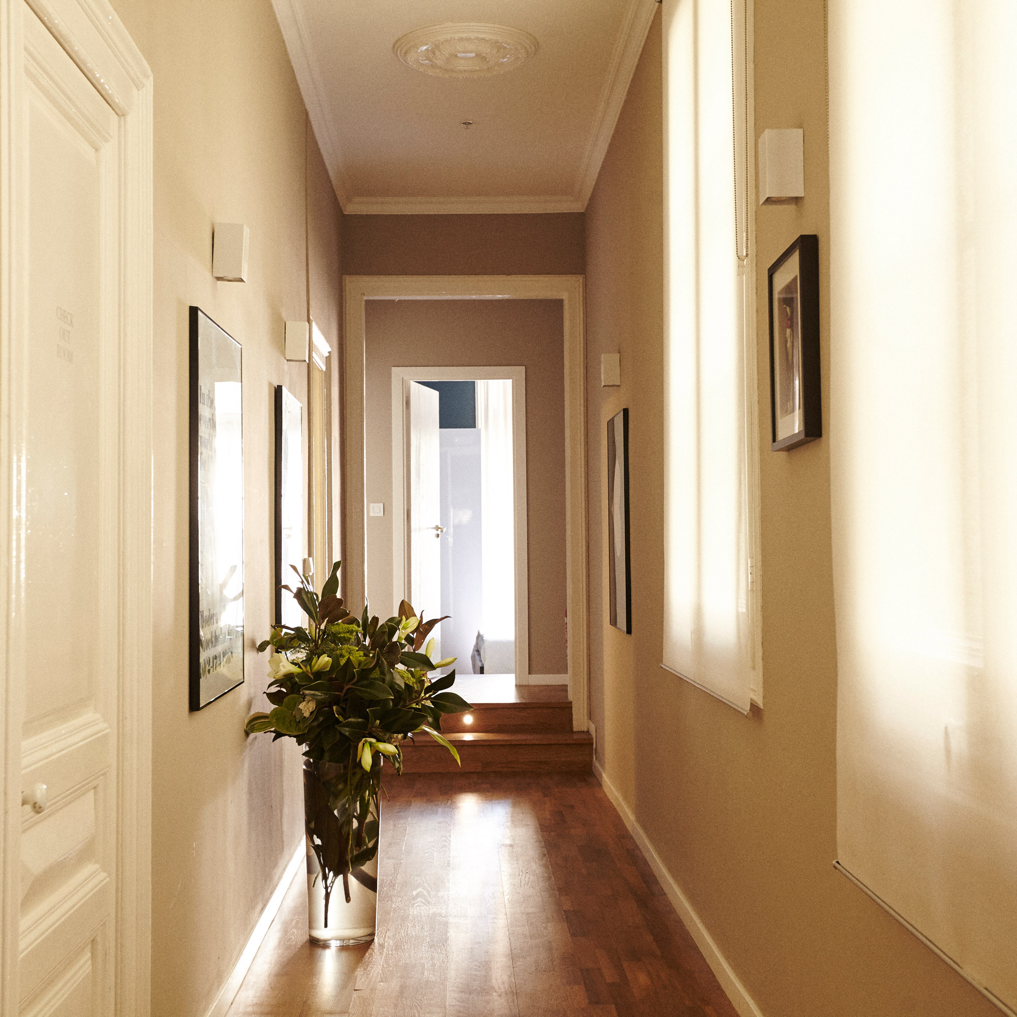 Couloir de style moderne dans des couleurs claires avec parquet au sol décoré de cadres au murs, avec un grand vase fleuri posé à même le sol.