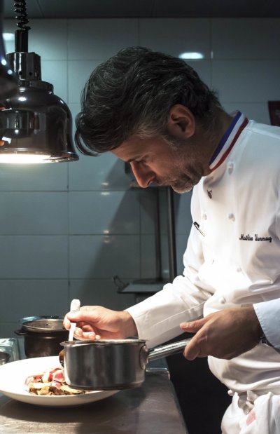Chef en veste de cuisinier avec col bleu blanc rouge, dans sa cuisine finalisant une assiette