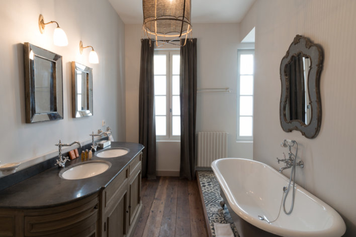 Salle de bain, baignoire sur pieds à droite face à laquelle se trouve une double vasque, en fond une fenêtre et longs rideaux