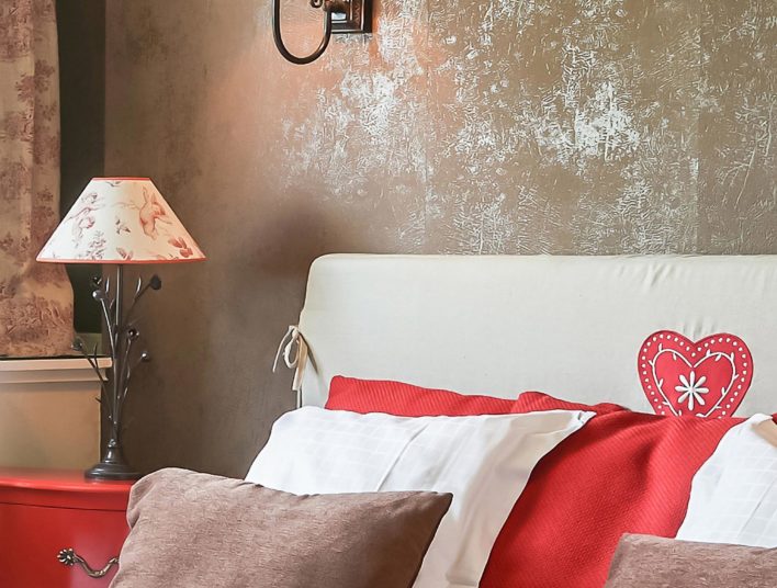 Détail décoration tête de lit couleur lin, coussin rouge, marron et blanc, deux lampes de chevets dont une applique