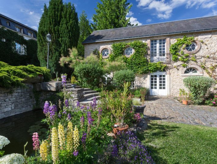 Longère recouverte de lierre grimpant, allée pavé et végétation diverse fleurie