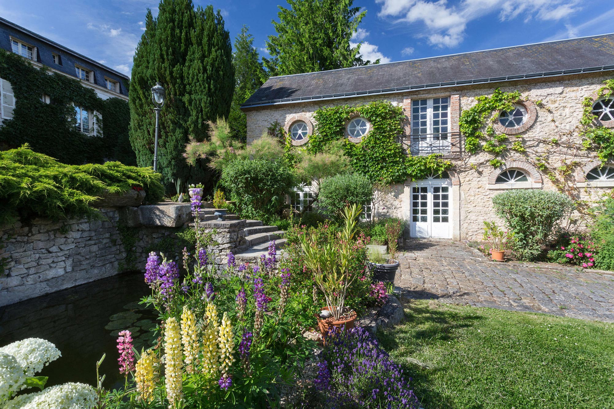 Longère recouverte de lierre grimpant, allée pavé et végétation diverse fleurie