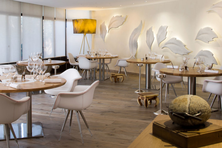Salle de restaurant avec chaise style scandinave, tables rondes et mur décoré de grandes feuilles blanche
