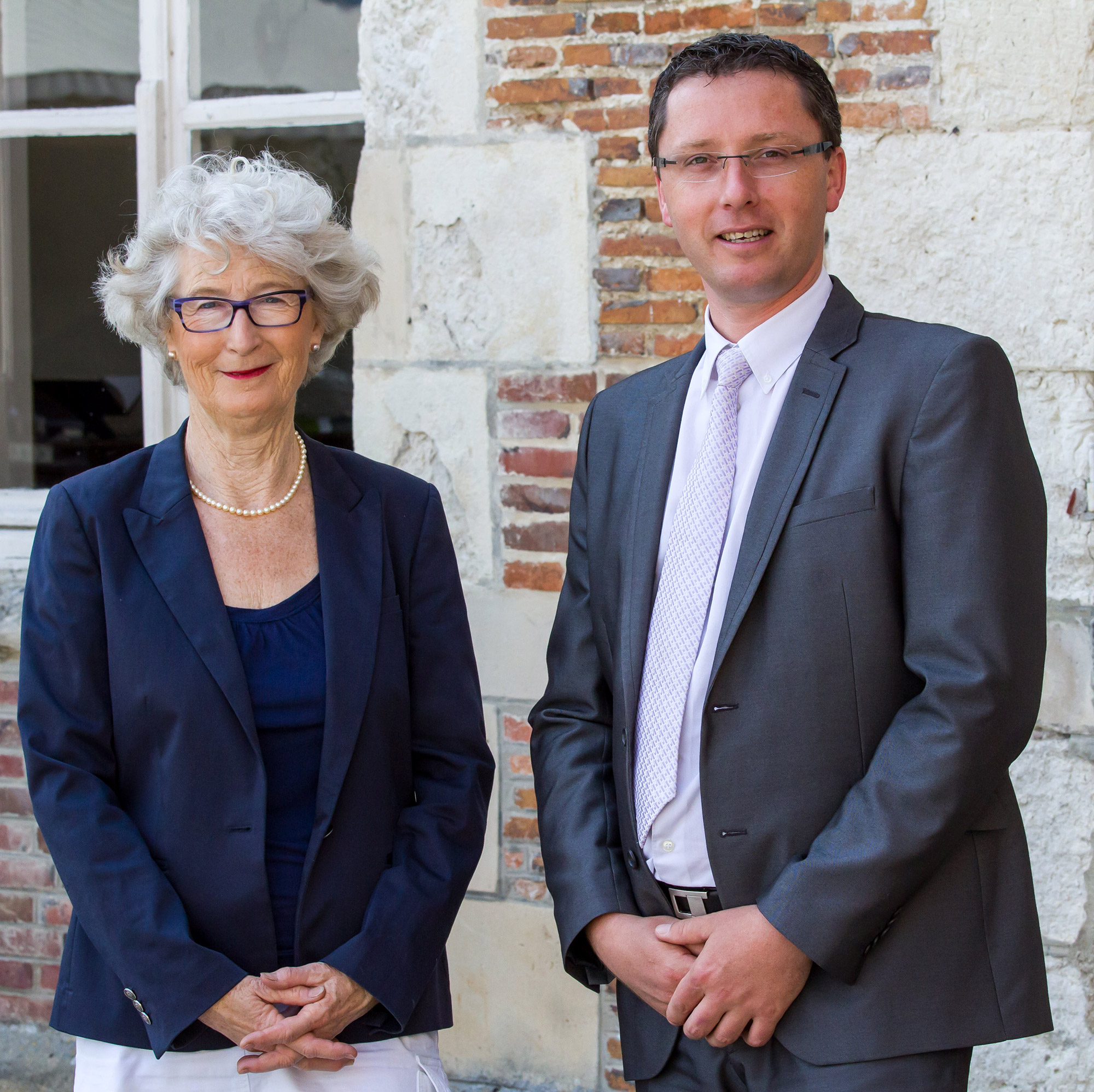 Photo d'une femme en blazer et pull bleu marine à côté d'un homme en, blazer gris et chemise mauve