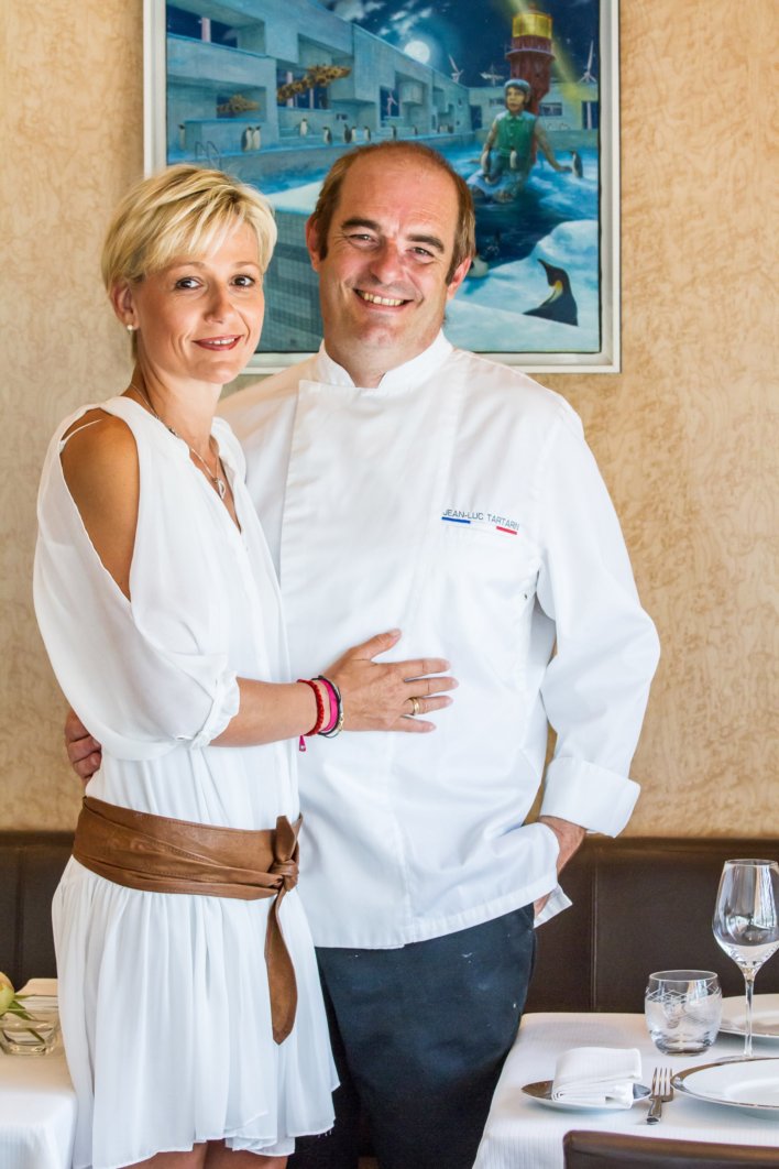 Annabelle habillé en blanc avec une ceinture marron qui est a coter de son mari avec sa main poser sur lui, et Jean-Luc Tartarin habillé de son tablier blanc et d'un pantalon qui à posé sa main derrière le dos de sa femme