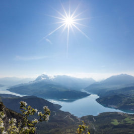 Plan panoramique sur des montagnes, une rivière, un grand ciel bleu et le soleil au centre de la photo