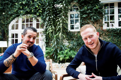 Photo de deux hommes assis en terrasse en pull et veste bleu, arrière plan mur de maison recouvert de lier