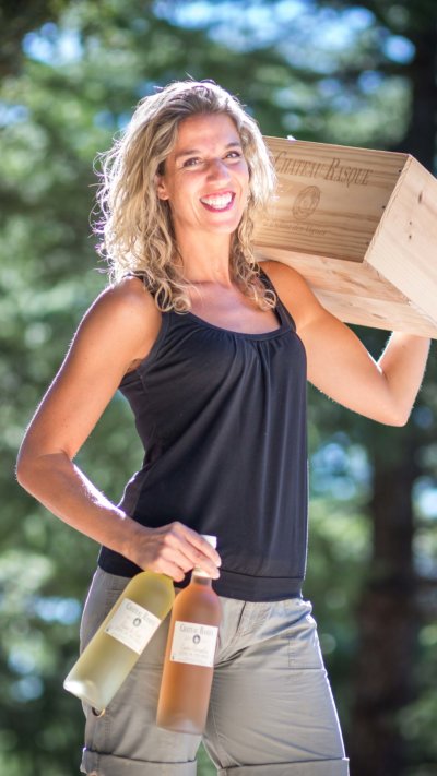 Femme tenant une caisse de vin sur l'épaule avec le bras gauche et deux bouteilles de vin (un blanc et un rosé) dans la main gauche
