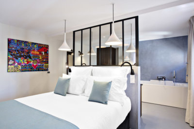 Chambre double avec tête de lit verrière donnant sur une salle de bain avec baignoire, une toile en peinture aérosol accroché sur le côté du lit