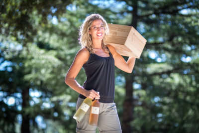 Femme tenant une caisse de vin sur l'épaule avec le bras gauche et deux bouteilles de vin (un blanc et un rosé) dans la main gauche