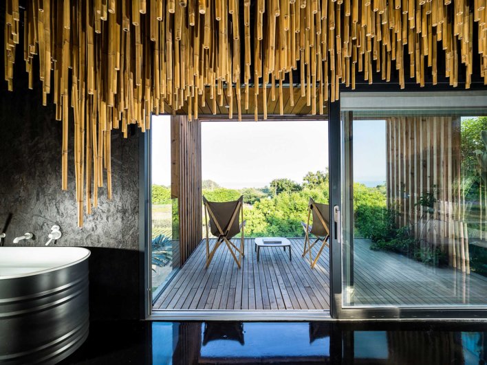 Baignoire dans salle de bain design avec mur noir en ardoise et bambou suspendu au dessus, vue sur terrasse privative et horizon verdoyant