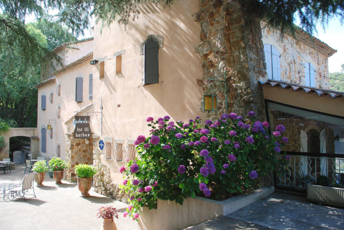 Photo de la façade prise de profil. On y voit des fleurs violettes au bord de la façades et quelques pots de fleurs à l'entrée et une chaise.