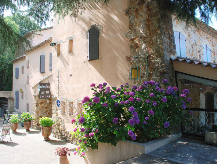 Photo de la façade prise de profil. On y voit des fleurs violettes au bord de la façades et quelques pots de fleurs à l'entrée et une chaise.