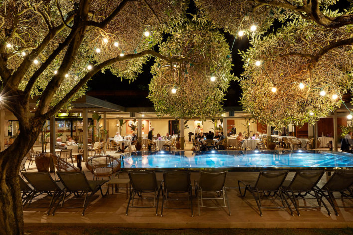 Photo de la piscine prise de nuit, on y voit autour des personnes se restaurées sur les tables qui sont en extérieures. Il y a des guirlande de lumières accrochées sur les arbres
