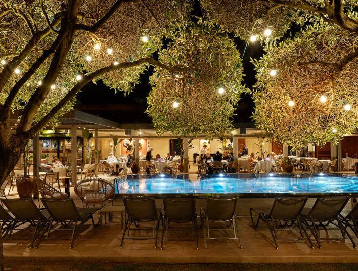 Photo de la piscine prise de nuit, on y voit autour des personnes se restaurées sur les tables qui sont en extérieures. Il y a des guirlande de lumières accrochées sur les arbres
