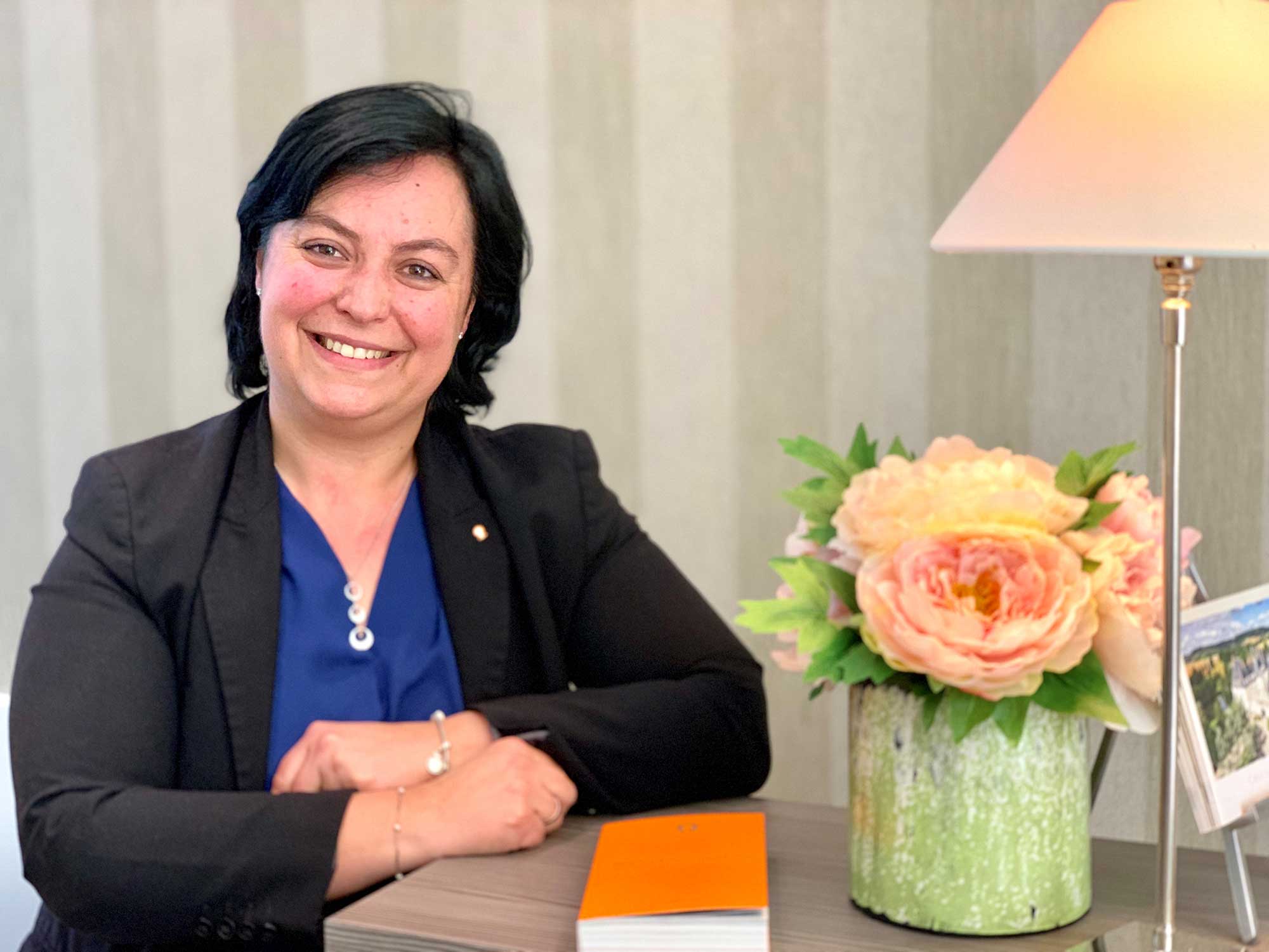 Femme accoudée à un bureau, blazer noir et chemisier bleu, un livre orange posé sur le bureau
