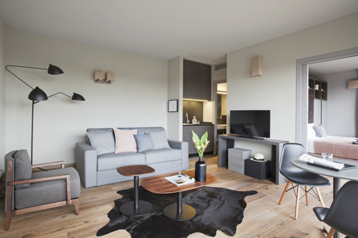 Photo de la suite double où l'on voit un salon avec une décoration design avec un canapé gris et des oreillers gris et rose, une petite table basse en bois et un meuble TV sur le coter. On aperçoit la cuisine et le lit.