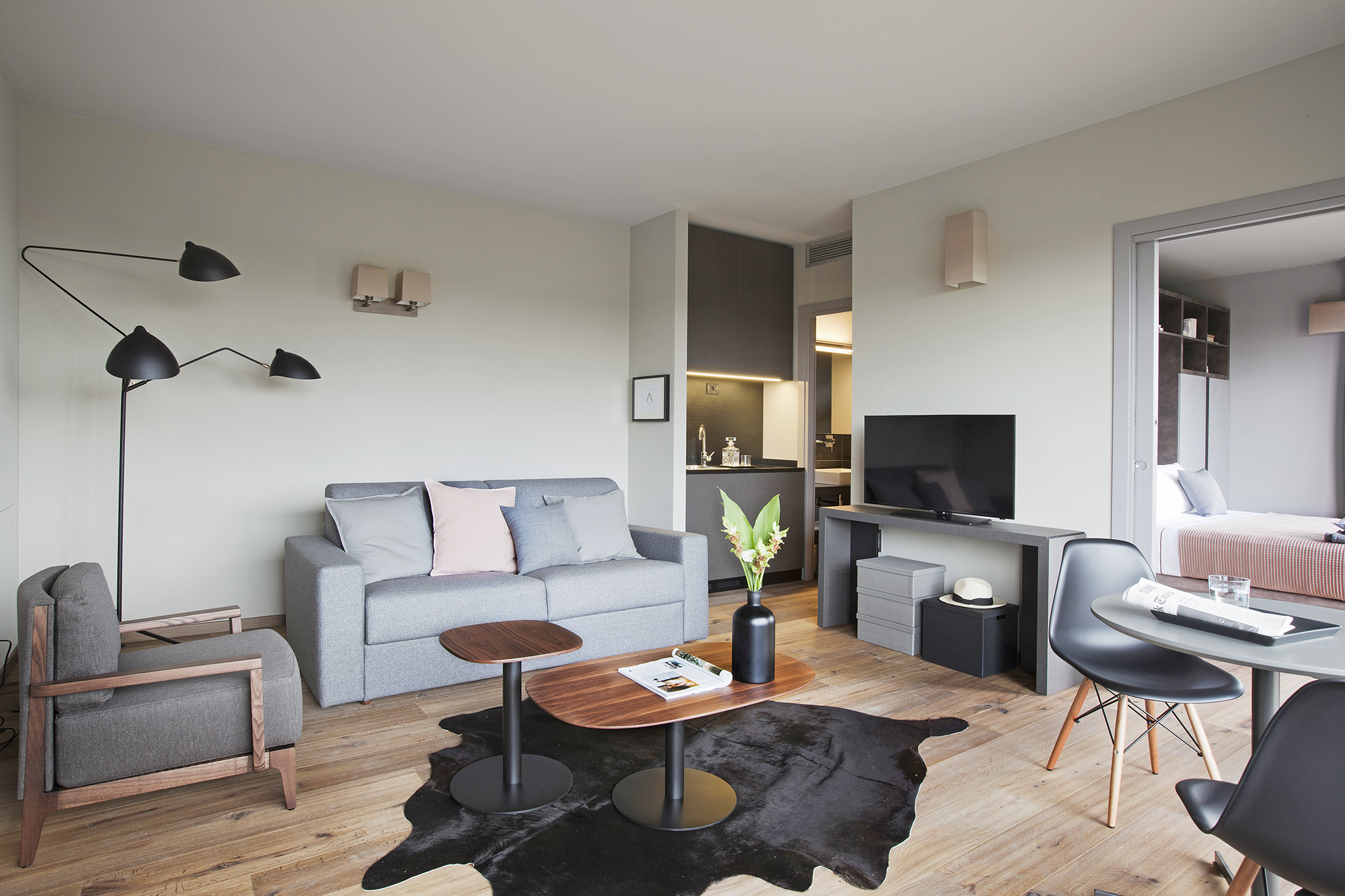 Photo de la suite double où l'on voit un salon avec une décoration design avec un canapé gris et des oreillers gris et rose, une petite table basse en bois et un meuble TV sur le coter. On aperçoit la cuisine et le lit.