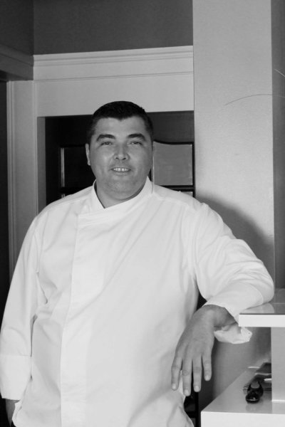 Plan américain noir et blanc d'un chef en veste de cuisine blanche, accoudée à un bar