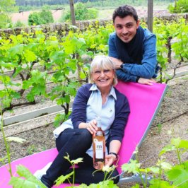 Femme en chemise bleu ciel dans un transat au coeur des vignes et un hommes derrière elle accoudé au transat, en chemise bleu électrique