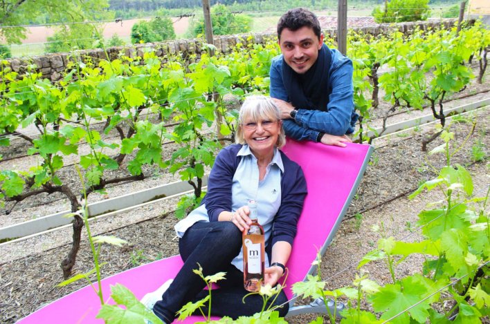 Femme en chemise bleu ciel dans un transat au coeur des vignes et un hommes derrière elle accoudé au transat, en chemise bleu électrique