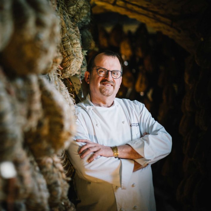 Photo portrait d'un chef en veste blanche, il pose devant des Culatello di Zibello, un jambon italien