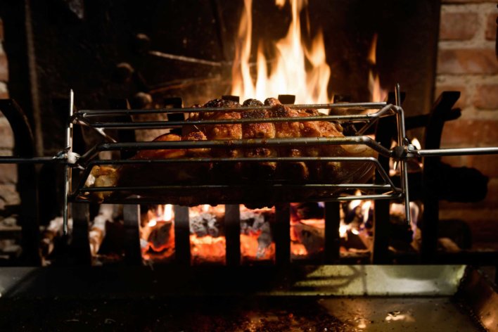 Pièce de viande à la broche qui tourne devant une cheminée au feu de bois