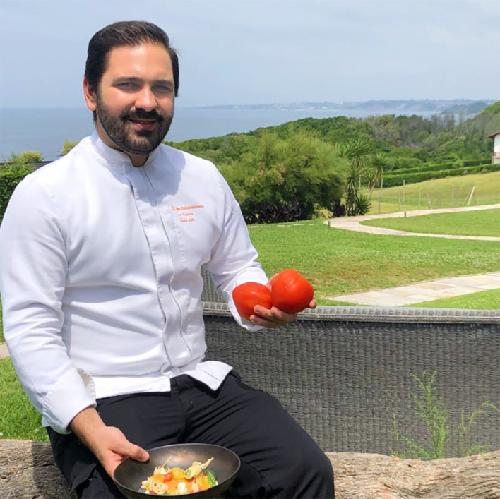 Chef en veste blanche tenant deux tomates rouges dans la main droite et une assiette dressée dans la main gauche, il est assis sur un muret avec vue mer