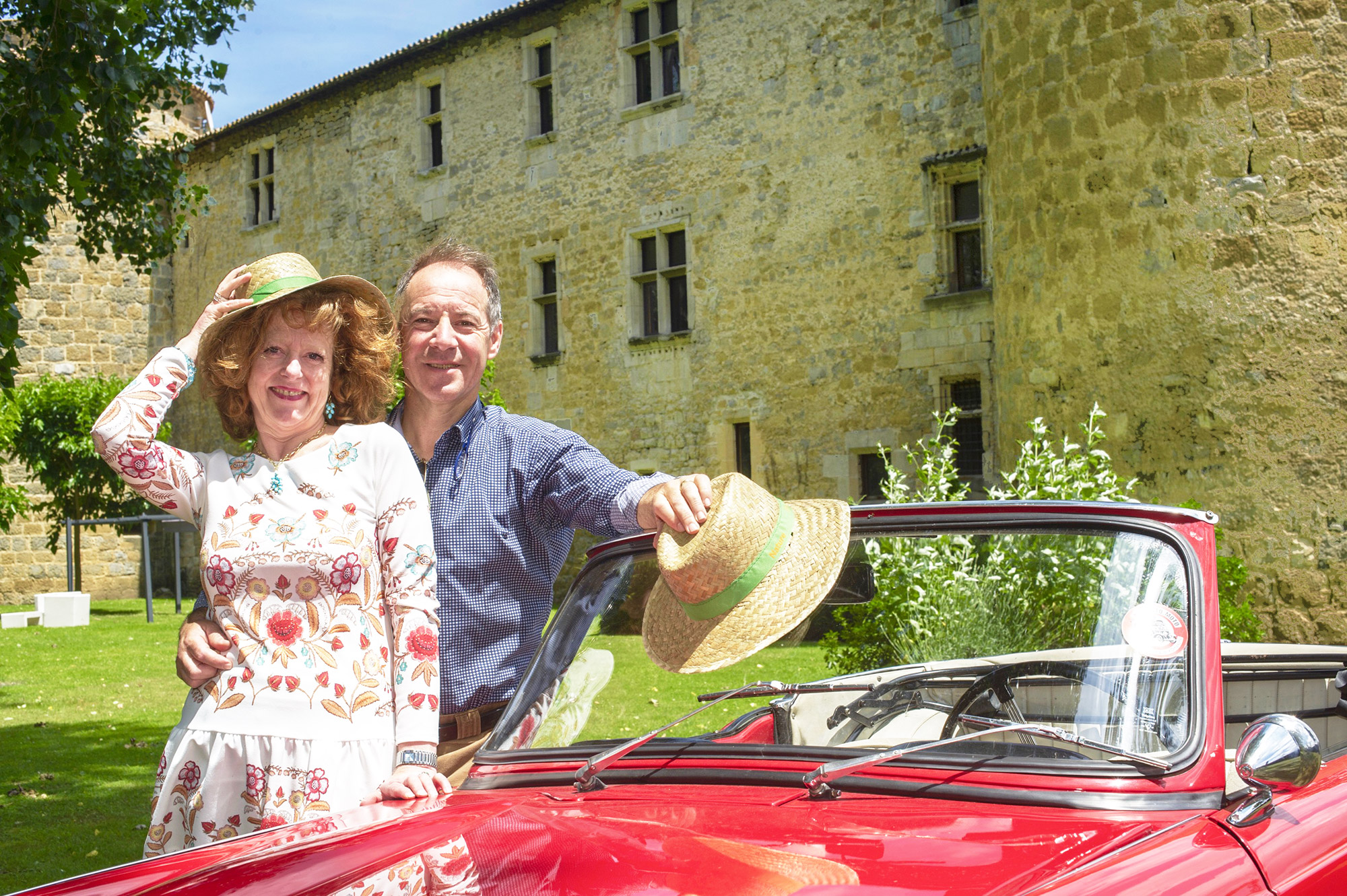 Une femme et un homme devant une voiture rouge avec un château en arrière-plan