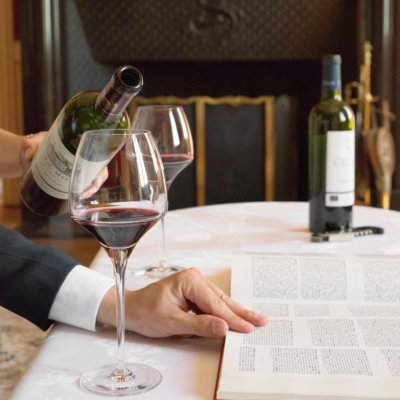 Vin servit au verre sur une table nappée blanche, un homme pose sa main droite sur un livre ouvert