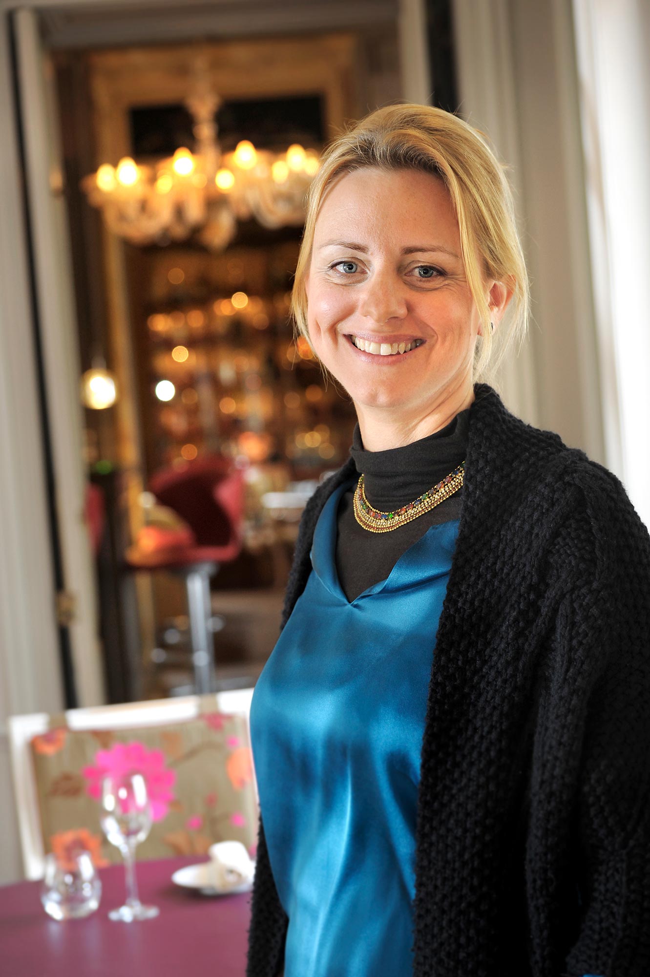 Plan américain d'une femme en gilet noir, top bleu et collier doré, arrière plan flouté salle de réception