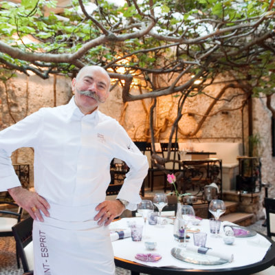 Chef en veste blanche, dans une cours intérieure ornée d'un arbre au dessus des tables dressées