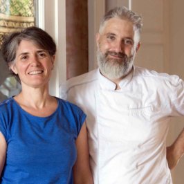 une femme en top bleu et un homme en veste de cuisine blanche l'un à côté de l'autre dans un salon