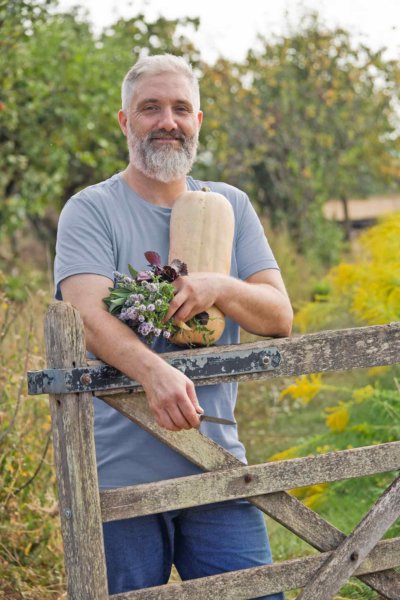 Homme appuyé sur une barrière en bois, tient dans ses bras une courge et des plantes aromatiques