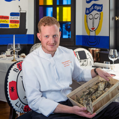Chef en veste de cuisine blanche, assis sur une chaise, tient une bourriche d'huître