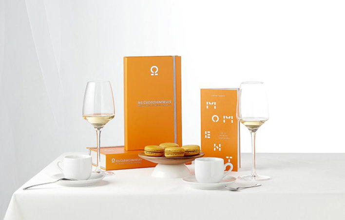 Coffret sur une table nappée d'une nappe de couleur blanche avec deux verres de vin blanc, deux petites tasses de café ainsi qu'une petite assiette de trois macarons