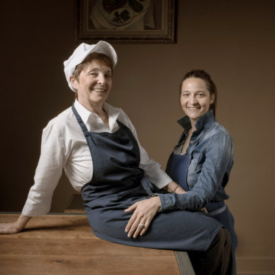 Deux femmes l'une à côté de l'autre, la première à gauche assises sur une table en tablier bleue et casquette blanche, la seconde assises à ses côtés en tablier bleu
