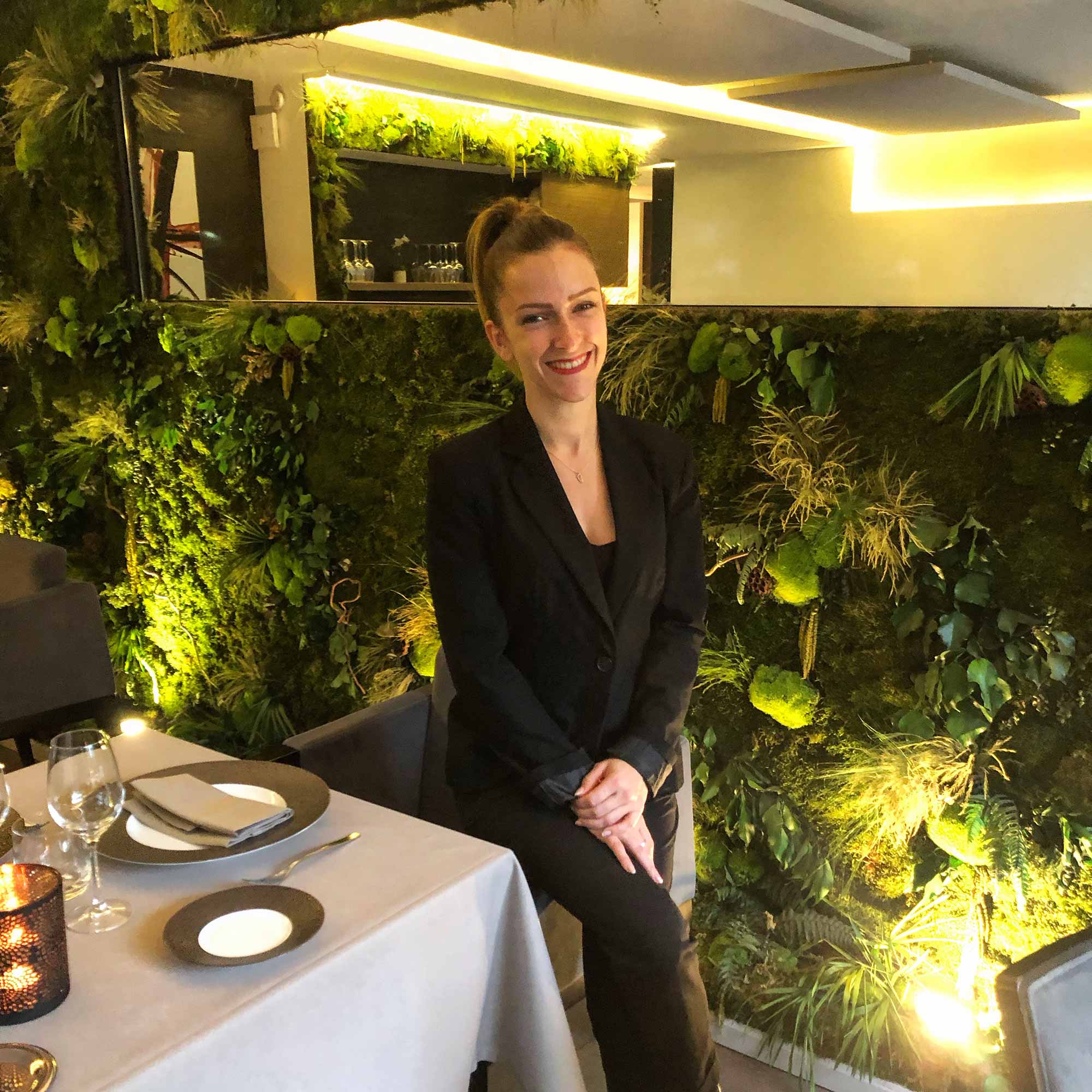 Femme vêtue de noir, dans une salle de restaurant avec un mur végétal en arrière-plan