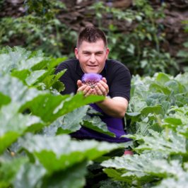 Photo portrait d'un chef, dans un potager, il tient un artichaud en fleur dans la main